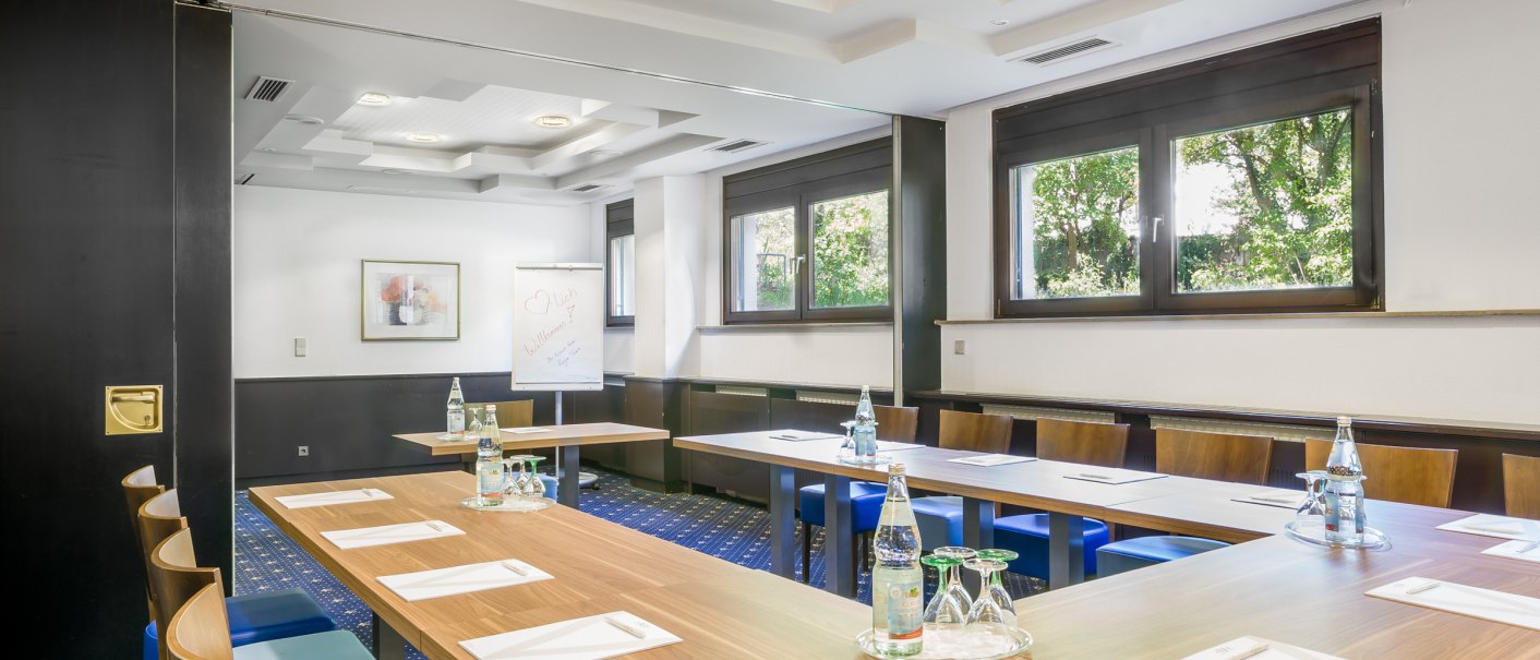 Meeting room with U-shaped table arrangement, water bottles and writing materials. Large windows and a flipchart in the background., © TOMAS
