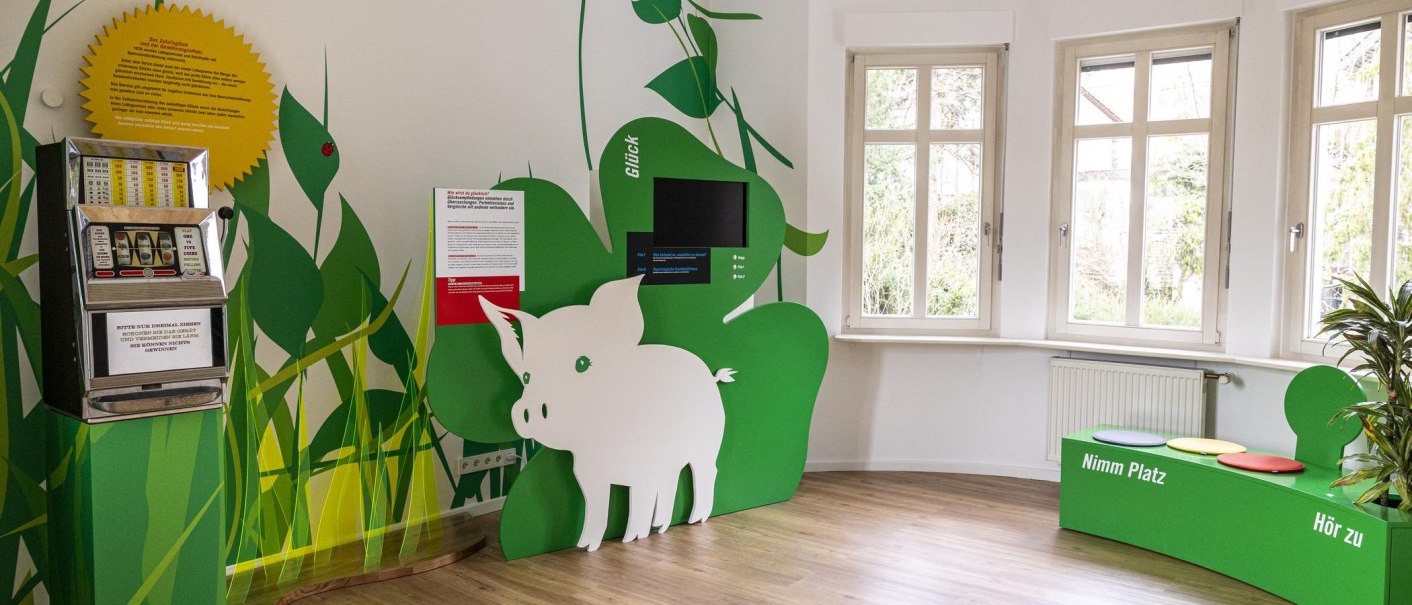 Ein Ausstellungsraum mit einem Spielautomaten, gr&uuml;ner Dekoration und Sitzgelegenheiten. Ein Schwein als Dekorationselement ist sichtbar., &copy; Stuttgart-Marketing GmbH, Sarah Schmid