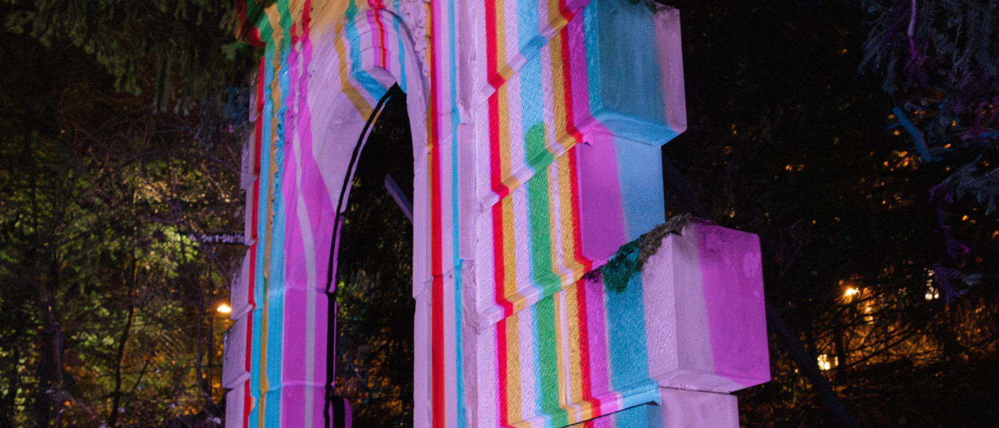 Ein Torbogen im Freien, beleuchtet mit bunten Lichtern in Pink, Blau und Gelb, umgeben von Bäumen bei Nacht., © Lichtgestalten