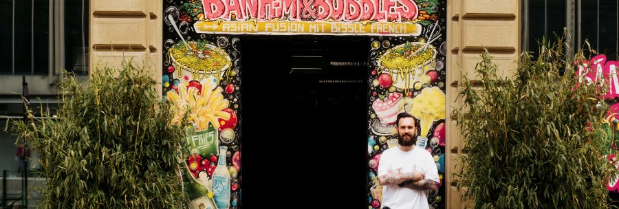 A man stands in front of the entrance to 'Banh Mi & Bubbles', surrounded by colorful graffiti and plants., © Banh Mi & Bubbles Restaurant / Bar