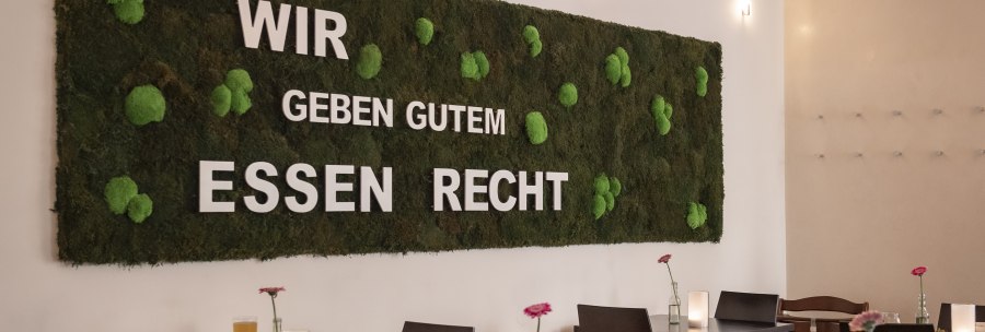 Restaurant mit Mooswand und dem Schriftzug 'Wir geben gutem Essen Recht'. Dunkle Tische, Kerzen und Blumen dekorieren den Raum., &copy; Speisekammer West