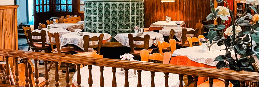 Cozy dining room with wooden furniture, a green tiled stove and decorative tablecloths. Hanging lamps and flower arrangements complete the ambience., © Weinstube Kachelofen