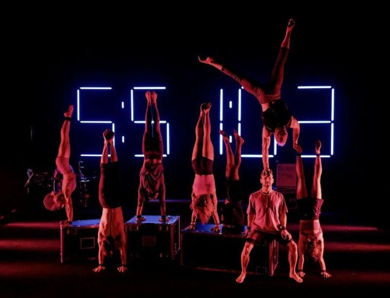 Acrobats perform impressive handstands in front of a large, glowing digital clock. The stage is kept in dark tones., &copy; Theaterhaus Stuttgart e.V.