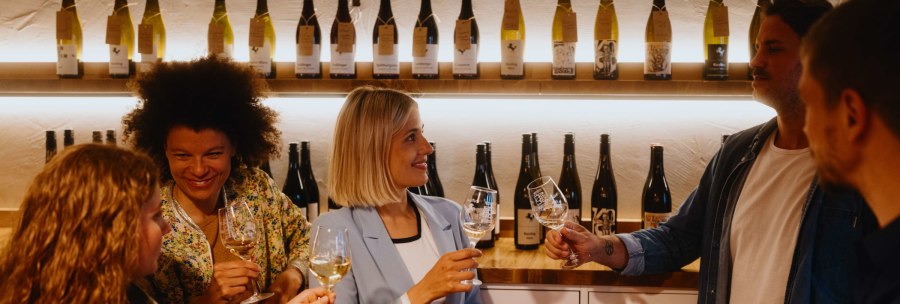 People in a wine shop clink glasses of wine. Wine bottles can be seen on shelves in the background., &copy; SMG, Alwin Maigler