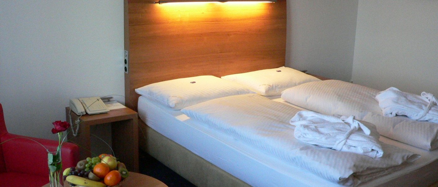 A cozy hotel room with double bed, bedside table, telephone, fruit basket and red rose on a table. Two bathrobes lie on the bed., © Hotel Fortuna