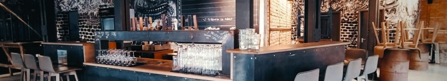 Modern bar with industrial design, high stools and wall art. Glasses are placed on the counter, in the background a table with drinks., © Kraftpaule Modern bar with industrial design, high stools and wall art. Glasses are placed on the counter, in the background a table with drinks., © Kraftpaule