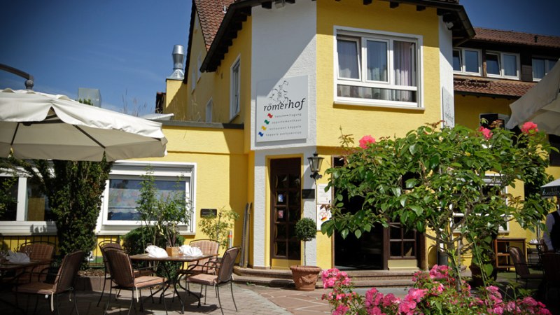 Gelbes Geb&auml;ude mit Terrasse, Tischen und St&uuml;hlen. Sonnenschirme und bl&uuml;hende Pflanzen im Vordergrund. Schild mit der Aufschrift 'r&ouml;merhof' an der Fassade., &copy; R&ouml;merhof Hotelbetriebs GmbH