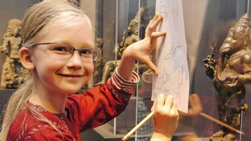 Ein Kind zeichnet in einem Museum neben einer südasiatischen Statue. Es trägt eine Brille und ein rotes Oberteil., © Linden-Museum Stuttgart Ein Kind zeichnet in einem Museum neben einer südasiatischen Statue. Es trägt eine Brille und ein rotes Oberteil., © Linden-Museum Stuttgart