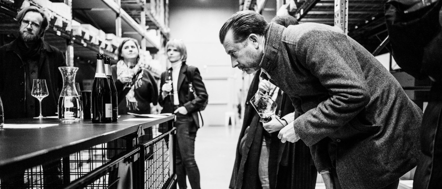 Eine Gruppe von Menschen steht in einem Lagerhaus und verkostet Wein. Ein Mann beugt sich vor, um ein Glas zu betrachten. Schwarz-Weiß-Fotografie., © club traube Eine Gruppe von Menschen steht in einem Lagerhaus und verkostet Wein. Ein Mann beugt sich vor, um ein Glas zu betrachten. Schwarz-Weiß-Fotografie., © club traube