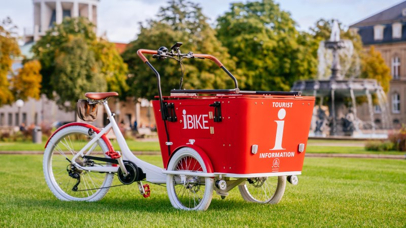 A red cargo bike with the inscription 'I-Bike' is parked on a meadow in front of a fountain and a building with a cupola., © Stuttgart-Marketing GmbH, Thomas Niedermüller