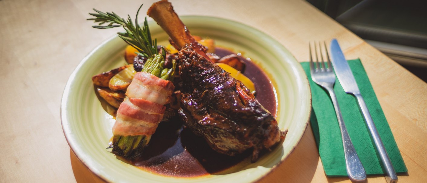 A plate with leg of lamb, asparagus wrapped in bacon and potatoes in sauce. Next to it are cutlery and a green napkin., © Rotenberger Weingärtle, Frederik Garlin