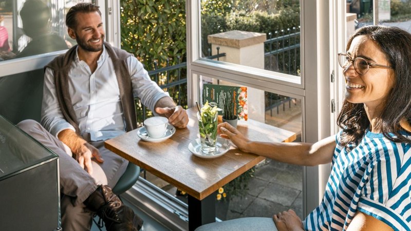 Zwei Personen sitzen in einem Café am Fenster, trinken Kaffee und unterhalten sich lächelnd. Sonnenlicht fällt durch das Fenster., © Stuttgart-Marketing GmbH, Sarah Schmid