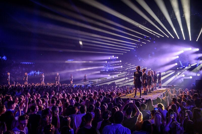 Konzertszene mit lila Lichtern, Models auf einem Laufsteg und einer großen Menschenmenge. Strahlenförmige Beleuchtung erzeugt eine dynamische Atmosphäre., © Fratelli BV Konzertszene mit lila Lichtern, Models auf einem Laufsteg und einer großen Menschenmenge. Strahlenförmige Beleuchtung erzeugt eine dynamische Atmosphäre., © Fratelli BV