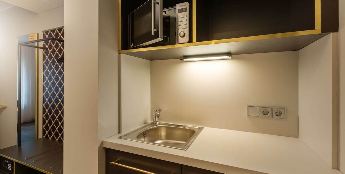 Kompakte Küche mit Spüle, Mikrowelle und Steckdosen. Dunkle Schränke mit goldenen Akzenten und Beleuchtung über der Arbeitsfläche., © Novum Hospitality