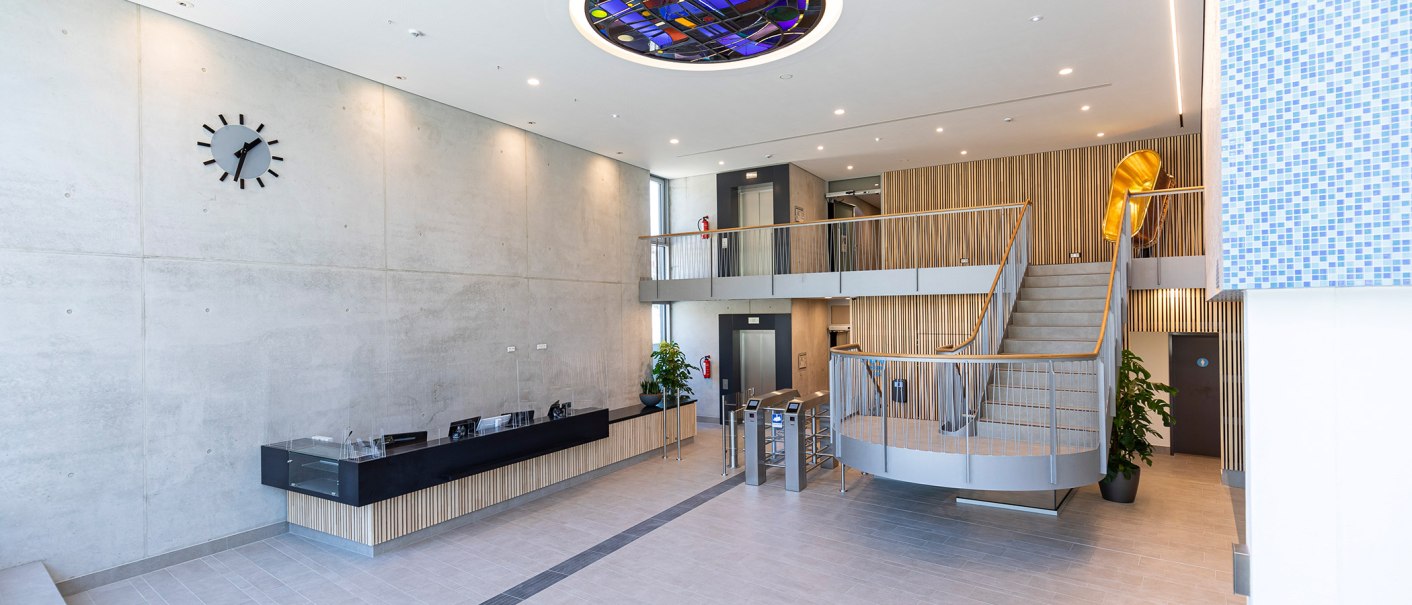 Modern foyer with reception desk, staircase, elevators and a large wall clock. The ceiling has a colorful glass element., © Stuttgarter Bäder