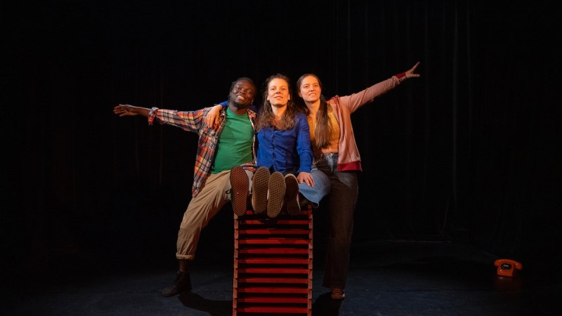 Drei Personen posieren fröhlich auf einer Bühne, zwei sitzen auf einem roten Kasten, eine steht daneben. Im Hintergrund ist ein schwarzer Vorhang., © Fotocredit Bettina Meister Drei Personen posieren fröhlich auf einer Bühne, zwei sitzen auf einem roten Kasten, eine steht daneben. Im Hintergrund ist ein schwarzer Vorhang., © Fotocredit Bettina Meister