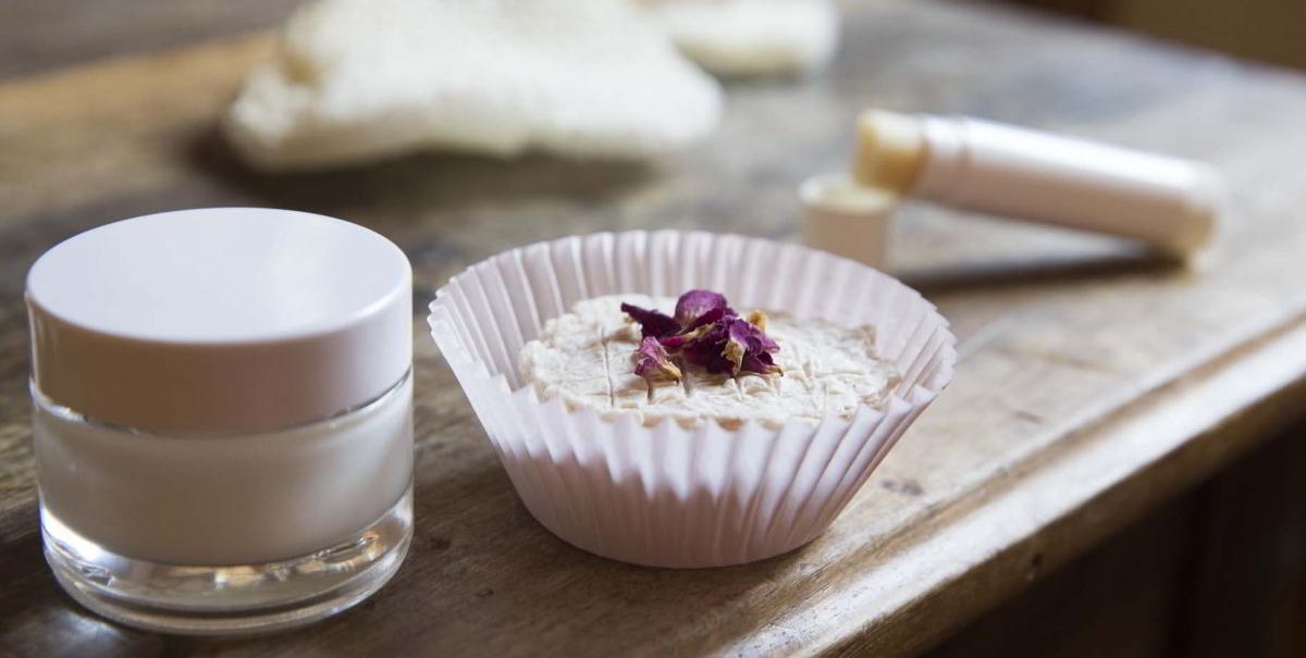 Ein Glas mit Creme, ein festes Kosmetikprodukt in einem Papierförmchen und ein Lippenpflegestift auf einem Holztisch., © hoponopo GmbH Ein Glas mit Creme, ein festes Kosmetikprodukt in einem Papierförmchen und ein Lippenpflegestift auf einem Holztisch., © hoponopo GmbH