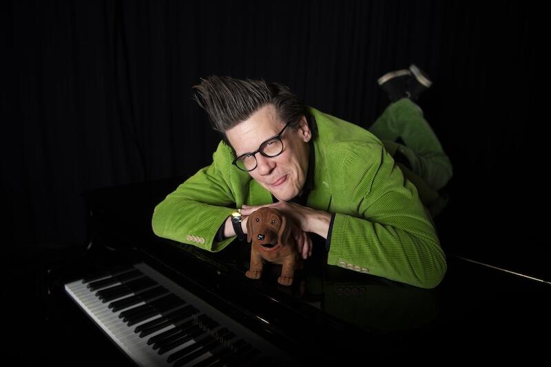 A person in a green suit is lying on a black piano, smiling and holding a dog toy. Dark background., © Renitenztheater Stuttgart e.V. A person in a green suit is lying on a black piano, smiling and holding a dog toy. Dark background., © Renitenztheater Stuttgart e.V.