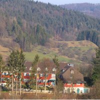 A red building with white windows stands in front of a wooded hill. Trees and bushes can be seen in the foreground., © DJH Baden-Württemberg e.V. A red building with white windows stands in front of a wooded hill. Trees and bushes can be seen in the foreground., © DJH Baden-Württemberg e.V.