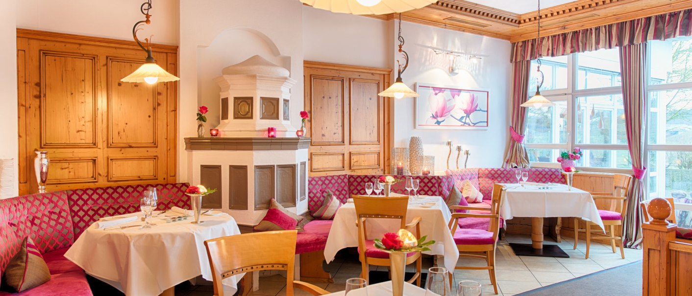Gemütliches Restaurant mit Holzmöbeln, rosa Polstern und Tischdecken. Ein Kamin und große Fenster sorgen für eine einladende Atmosphäre., © Landhaus Franz Feckl