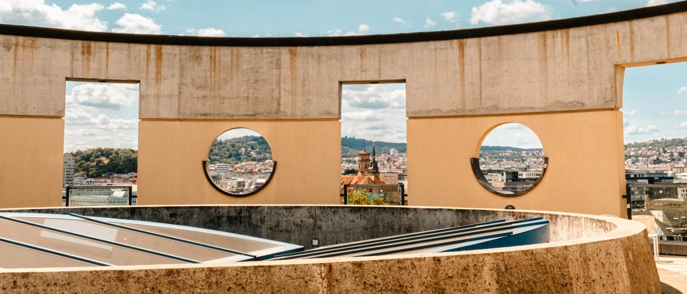 Betonwand mit runden und rechteckigen Öffnungen, durch die eine Stadtlandschaft mit Hügeln und Gebäuden sichtbar ist., © Stuttgart-Marketing GmbH, Sarah Schmid