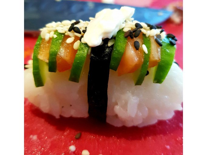 Sushi-Nigiri mit Lachs, Gurke, Sesam und Wasabi auf rotem Untergrund. Ein Streifen Nori hält die Zutaten zusammen., © Kultweine-Kochwelten-Kunstücke - Kochschule Sonja Lenz Sushi-Nigiri mit Lachs, Gurke, Sesam und Wasabi auf rotem Untergrund. Ein Streifen Nori hält die Zutaten zusammen., © Kultweine-Kochwelten-Kunstücke - Kochschule Sonja Lenz