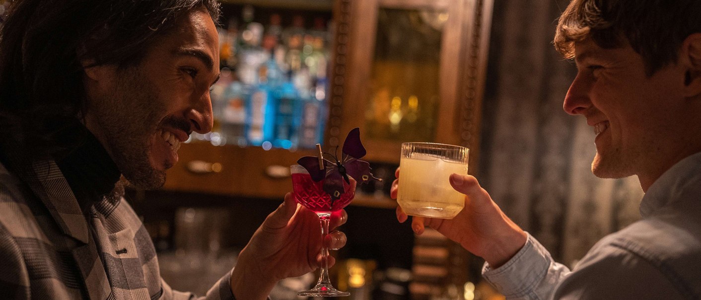 Zwei M&auml;nner sto&szlig;en in einer Bar mit Cocktails an, einer in einem karierten Sakko, der andere in einem hellen Hemd. Im Hintergrund sind Flaschen zu sehen., &copy; SMG, Martina Denker