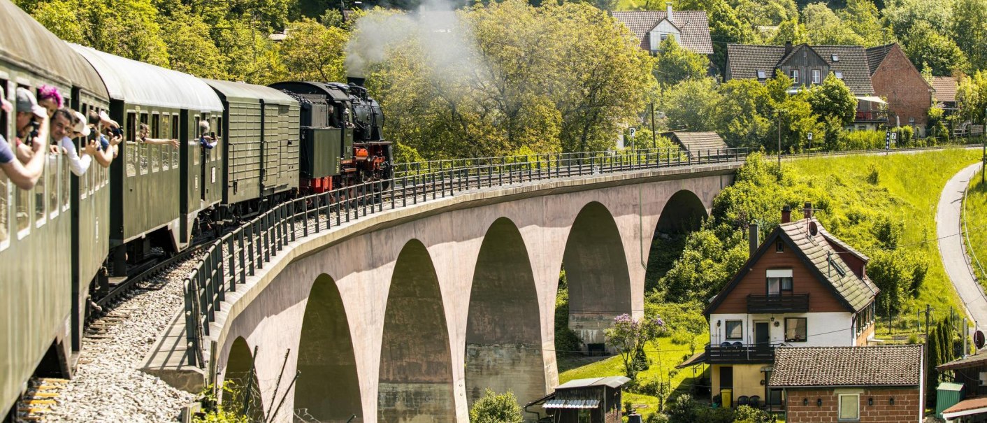 A steam locomotive pulls a train across a bridge in a green, rural landscape. People are looking out the windows and taking pictures., &copy; SMG Stuttgart Marketing GmbH - Sarah Schmid