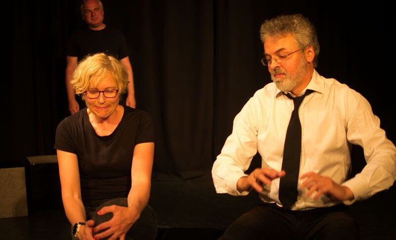 Zwei Personen sitzen auf einer Bühne, eine Frau und ein Mann, in einem dramatisch beleuchteten Raum. Eine weitere Person steht im Hintergrund., © Theater Atelier