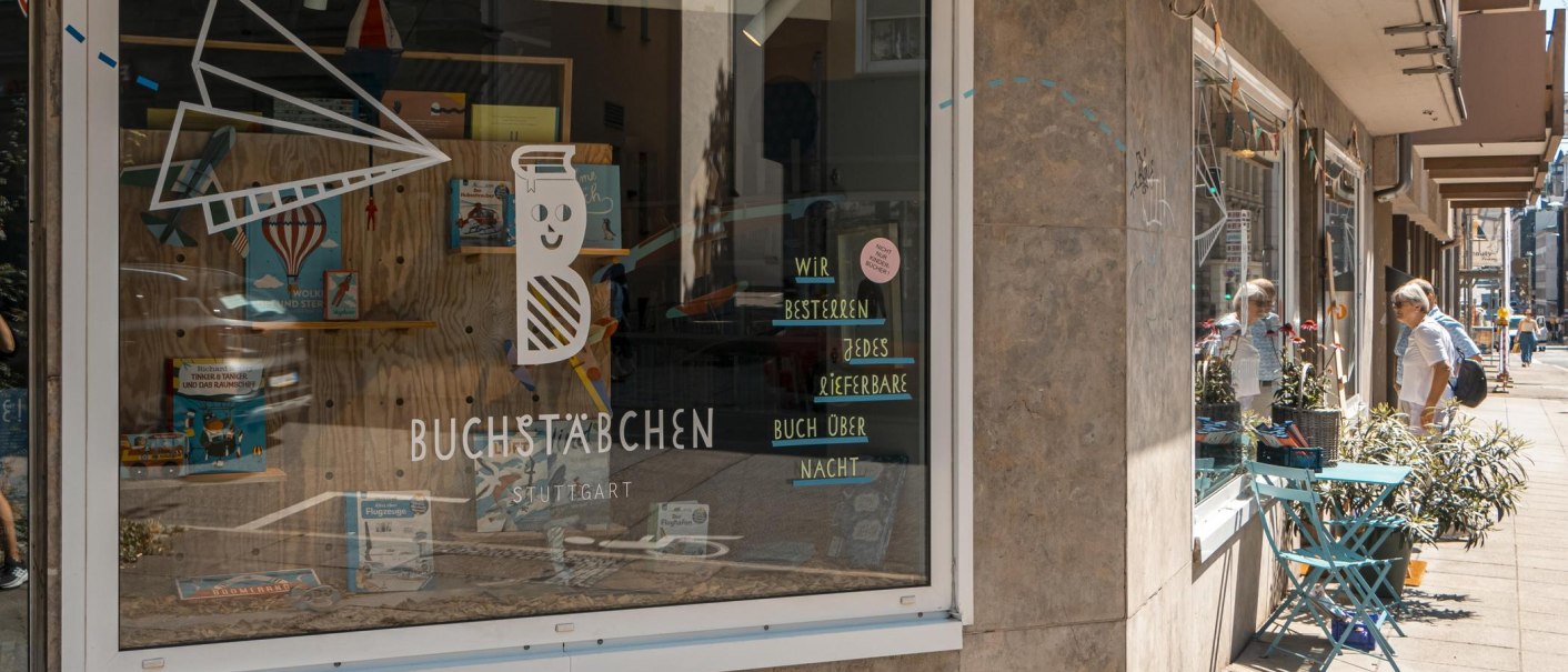 Schaufenster des Buchladens 'Buchstäbchen' in Stuttgart. Dekoriert mit Büchern und einem Papierflieger. Passanten betrachten das Schaufenster., © SMG, Sarah Schmid