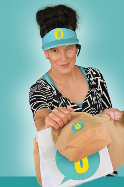 Frau mit blauem Visier und Headset reicht eine Papiert&uuml;te mit Logo vor t&uuml;rkisfarbenem Hintergrund., &copy; Renitenztheater Stuttgart e.V.