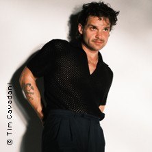 Ein Mann in einem schwarzen Hemd posiert vor einem weißen Hintergrund. Er hat lockiges Haar und trägt eine dunkle Hose., © links im Bild Ein Mann in einem schwarzen Hemd posiert vor einem weißen Hintergrund. Er hat lockiges Haar und trägt eine dunkle Hose., © links im Bild