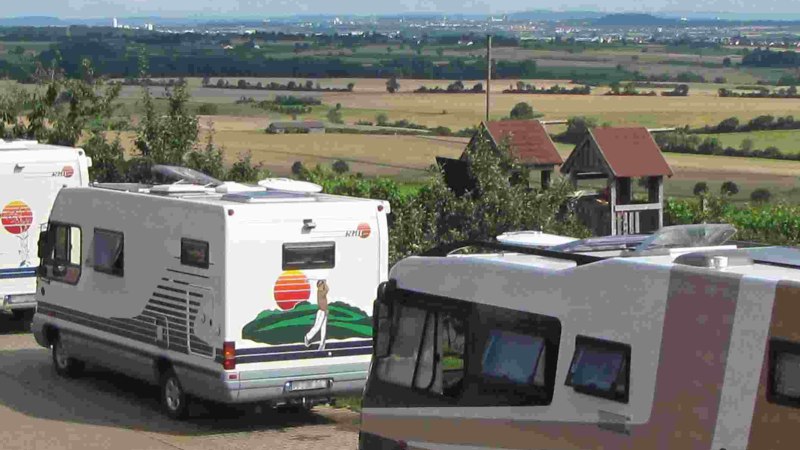 Wohnmobile parken auf einem Stellplatz mit Blick auf eine weite Landschaft und Felder. Im Hintergrund sind Hügel und eine Stadt zu sehen., © PANORAMAWEINGUT Baumgärtner Wohnmobile parken auf einem Stellplatz mit Blick auf eine weite Landschaft und Felder. Im Hintergrund sind Hügel und eine Stadt zu sehen., © PANORAMAWEINGUT Baumgärtner