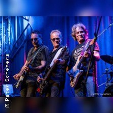 Drei Musiker performen mit Gitarren auf einer B&uuml;hne, umgeben von blauer Beleuchtung. Sie wirken konzentriert und energiegeladen., &copy; links im Bild