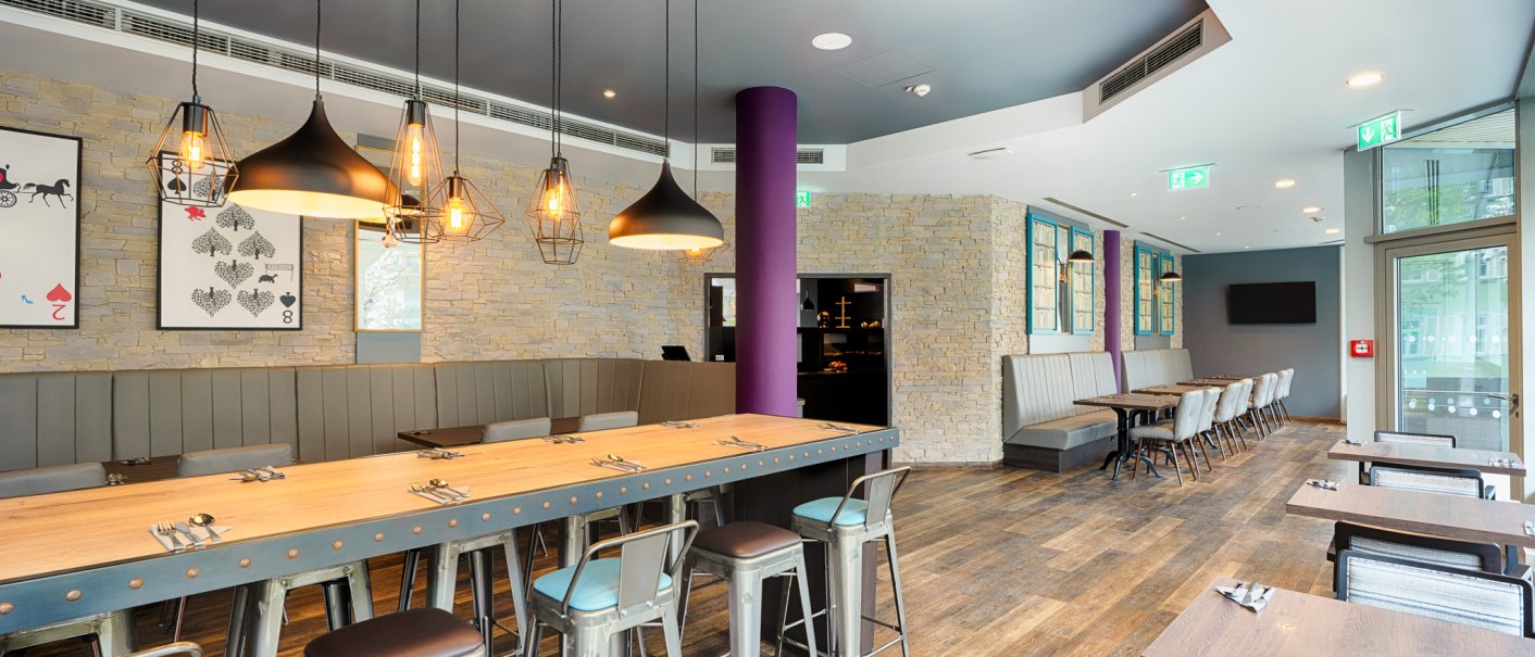 Moderner Fr&uuml;hst&uuml;cksbereich mit langen Holztischen, Metallst&uuml;hlen und stilvollen H&auml;ngelampen. Steinw&auml;nde und gro&szlig;e Fenster schaffen eine einladende Atmosph&auml;re., &copy; Premier Inn Germany