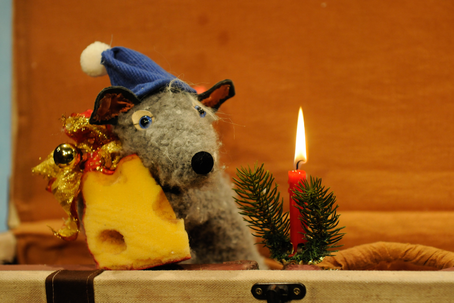Eine Stoffratte mit blauer Mütze hält ein Stück Käse. Daneben steht eine brennende rote Kerze mit Tannenzweig. Weihnachtliche Szene., © TheaterFusion