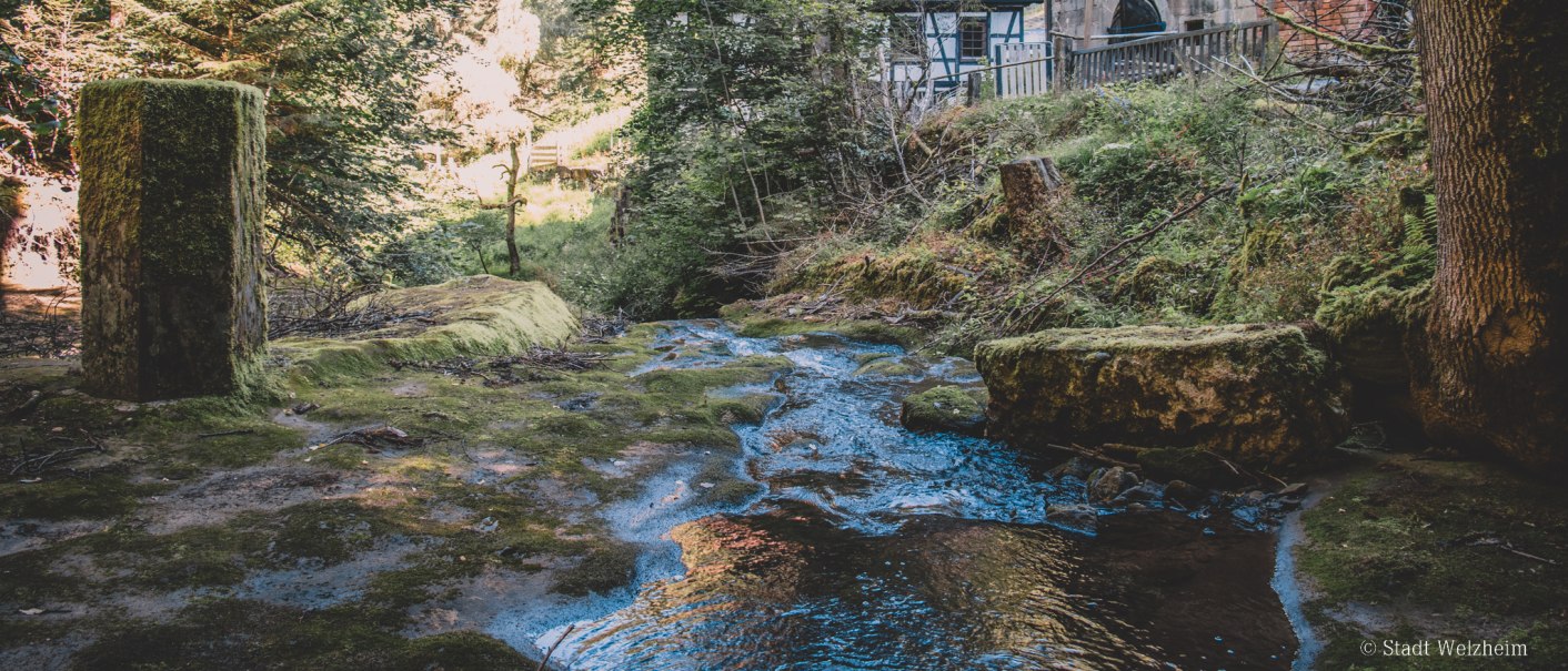 A stream flows through a forest, past an old half-timbered house. Moss-covered rocks and trees surround the scene., &copy; Stadt Welzheim