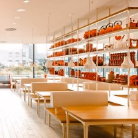 Modernes Restaurant mit weißen Tischen und Regalen voller roter Dekorationen. Große Fenster bieten Blick nach draußen., © Breuninger Modernes Restaurant mit weißen Tischen und Regalen voller roter Dekorationen. Große Fenster bieten Blick nach draußen., © Breuninger