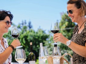 Zwei Frauen genießen Wein im Freien, stoßen mit Gläsern an. Im Hintergrund sind Bäume und ein blauer Himmel zu sehen., © SMG, Thomas Niedermüller