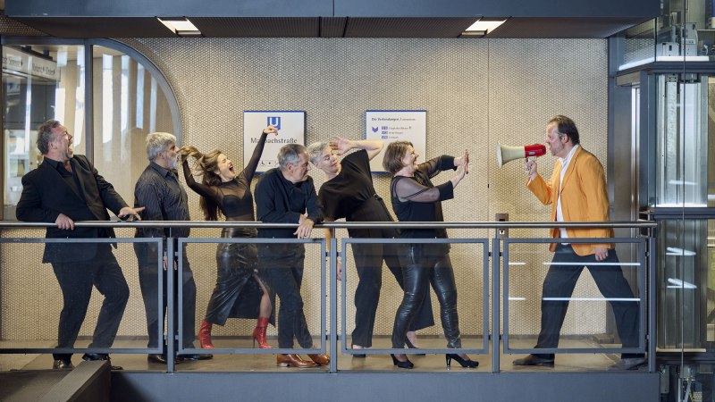 Eine Gruppe von Menschen steht auf einem Balkon. Eine Person im orangefarbenen Sakko spricht in ein Megafon, während die anderen reagieren., © Martin Sigmund Eine Gruppe von Menschen steht auf einem Balkon. Eine Person im orangefarbenen Sakko spricht in ein Megafon, während die anderen reagieren., © Martin Sigmund