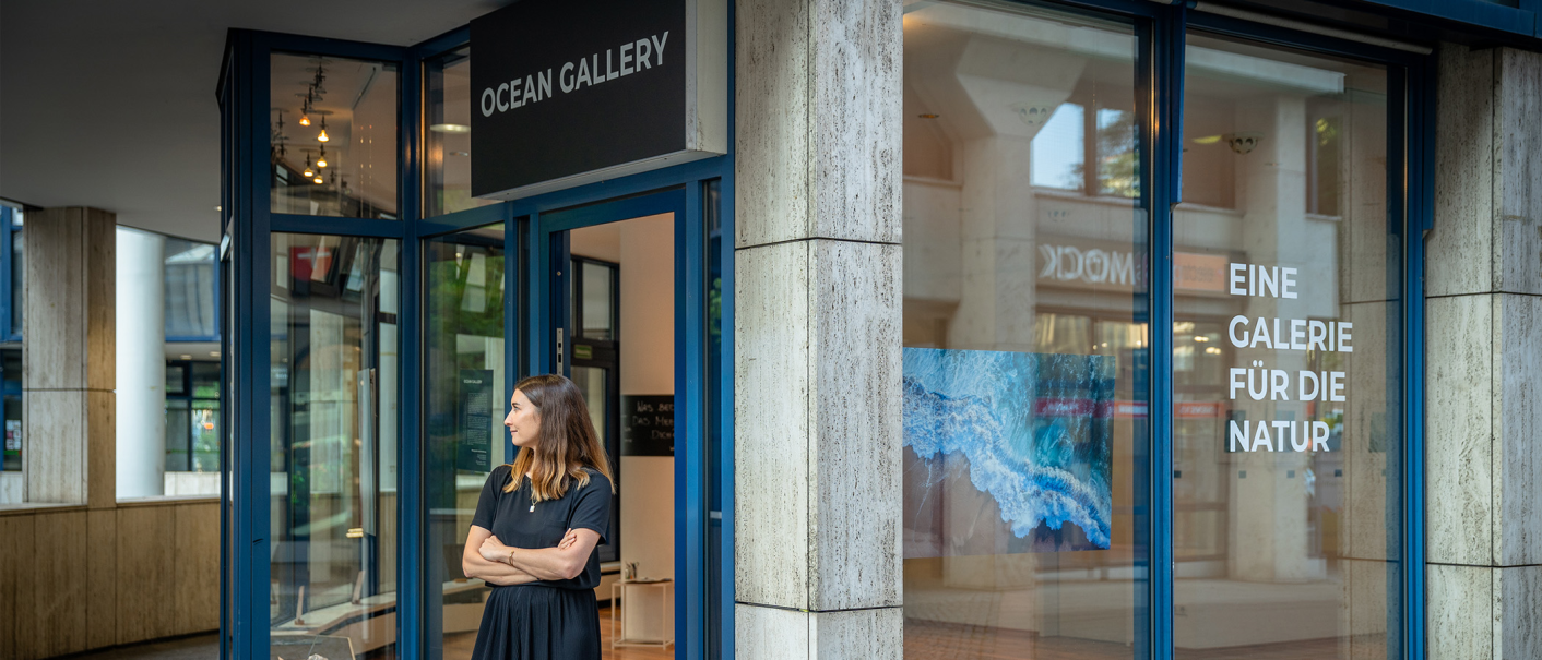 A woman stands in front of the Ocean Gallery. The shop window reads "A gallery for nature". The gallery has blue frames and a picture of nature in the window., © OCEAN GALLERY A woman stands in front of the Ocean Gallery. The shop window reads "A gallery for nature". The gallery has blue frames and a picture of nature in the window., © OCEAN GALLERY