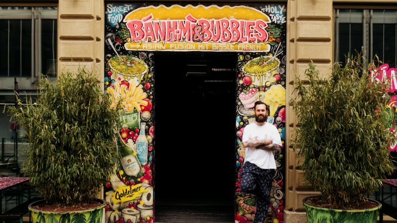 A man stands in front of the entrance to 'Banh Mi & Bubbles', surrounded by colorful graffiti and plants., &copy; Banh Mi & Bubbles Restaurant / Bar