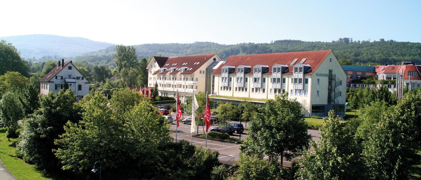 Geb&auml;udekomplex mit roten D&auml;chern, umgeben von B&auml;umen und H&uuml;geln in Bad Boll. Mehrere Flaggen sind vor dem Geb&auml;ude zu sehen., &copy; Seminaris Hotel Bad Boll