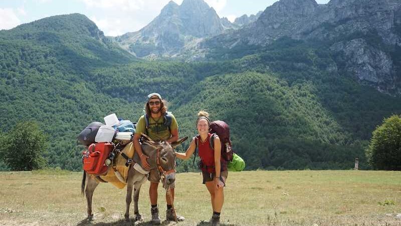Eine Person mit einem Esel, beide schwer bepackt, auf einer malerischen Bergstra&szlig;e. Die Umgebung ist von gr&uuml;nen W&auml;ldern und hohen Bergen gepr&auml;gt., &copy; Theaterhaus Stuttgart e.V.