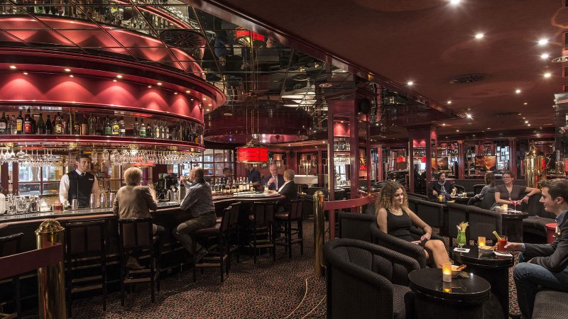 Elegante Bar mit rotem Dekor, Spiegeln und ged&auml;mpfter Beleuchtung. G&auml;ste sitzen an der Theke und an Tischen, w&auml;hrend ein Barkeeper Getr&auml;nke serviert., &copy; Maritim Hotels