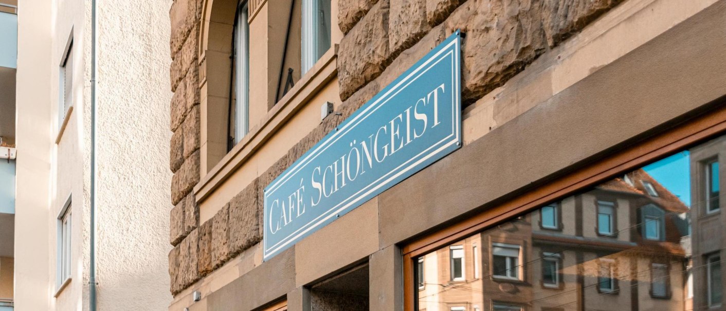 Das Caf&eacute; Sch&ouml;ngeist zeigt ein blaues Schild an einer historischen Steinwand. Im Fenster spiegelt sich die umliegende Architektur wider., &copy; SMG Stuttgart Marketing GmbH - Sarah Schmid