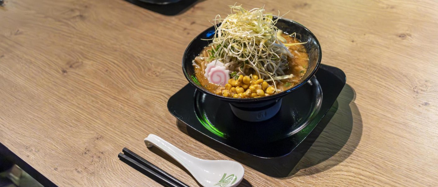 Eine Sch&uuml;ssel Ramen mit Mais, Narutomaki und Gem&uuml;se auf einem Holztisch. Daneben liegen Essst&auml;bchen und ein L&ouml;ffel., &copy; SMG, Sarah Schmid