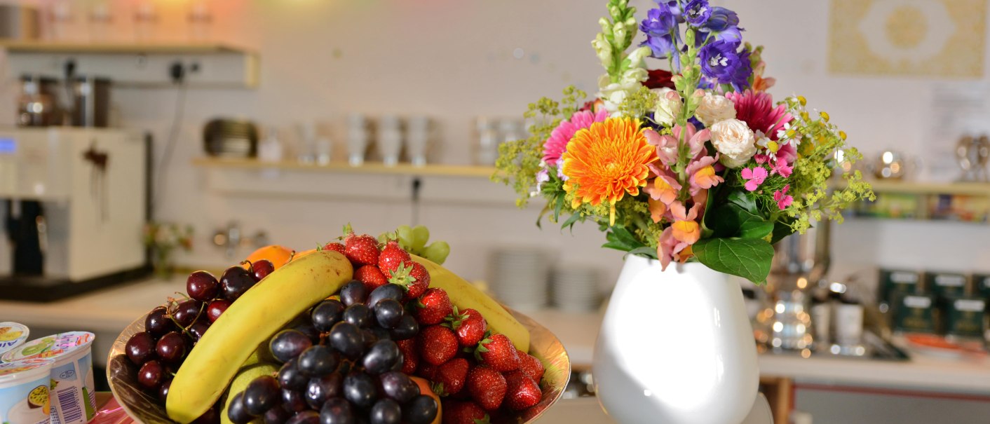 Ein Frühstücksbuffet mit einer Obstschale voller Bananen, Trauben und Erdbeeren neben einem bunten Blumenstrauß in einer weißen Vase., © Abalon Hotel Ideal Ein Frühstücksbuffet mit einer Obstschale voller Bananen, Trauben und Erdbeeren neben einem bunten Blumenstrauß in einer weißen Vase., © Abalon Hotel Ideal