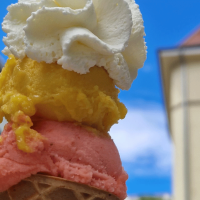 An ice cream cone with three scoops in different colors and whipped cream, against a background of buildings and blue sky., &copy; kesselglueck.de