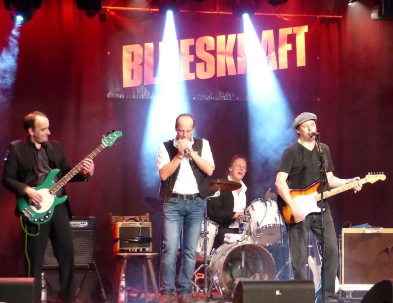 Eine Bluesband performt auf der B&uuml;hne. Der Bassist spielt eine gr&uuml;ne Gitarre, der S&auml;nger spielt Mundharmonika, der Schlagzeuger sitzt im Hintergrund und der Gitarrist singt., &copy; Veranstalter